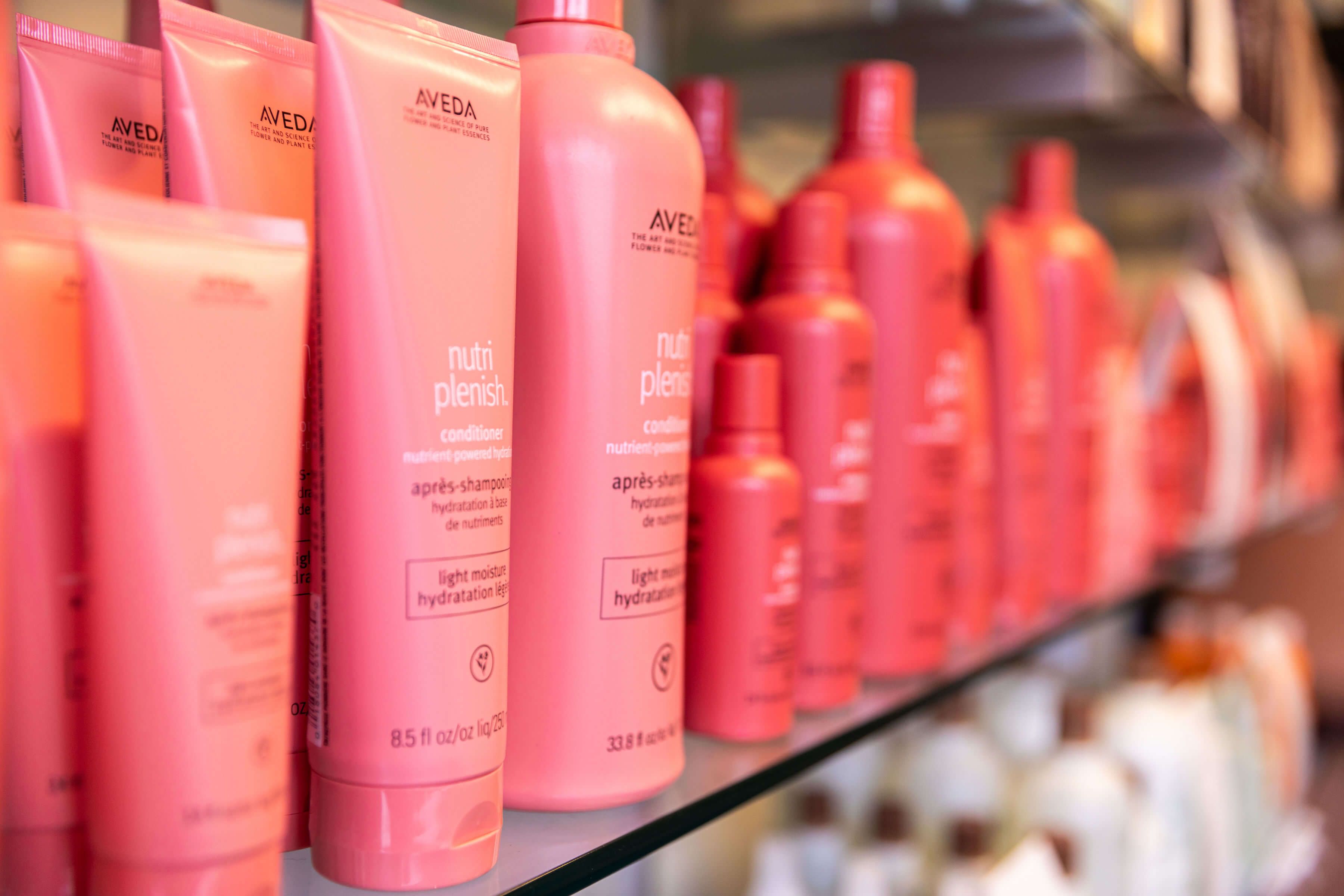 - Salone Estetica in Winchester, MA Close-up of several pink bottles and tubes of Aveda hair care products arranged on a glass shelf in an Aveda salon, perfect for hair stylists or those seeking keratin treatments. - Salone Estetica in Winchester, MA