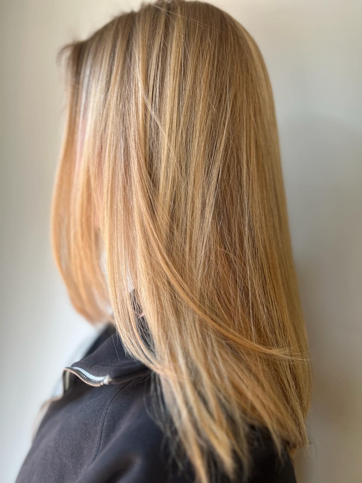 - Salone Estetica in Winchester, MA A person with straight, shoulder-length blonde hair is shown in profile, styled by a professional hair stylist at an Aveda salon, wearing a black top and standing against a plain light background. - Salone Estetica in Winchester, MA