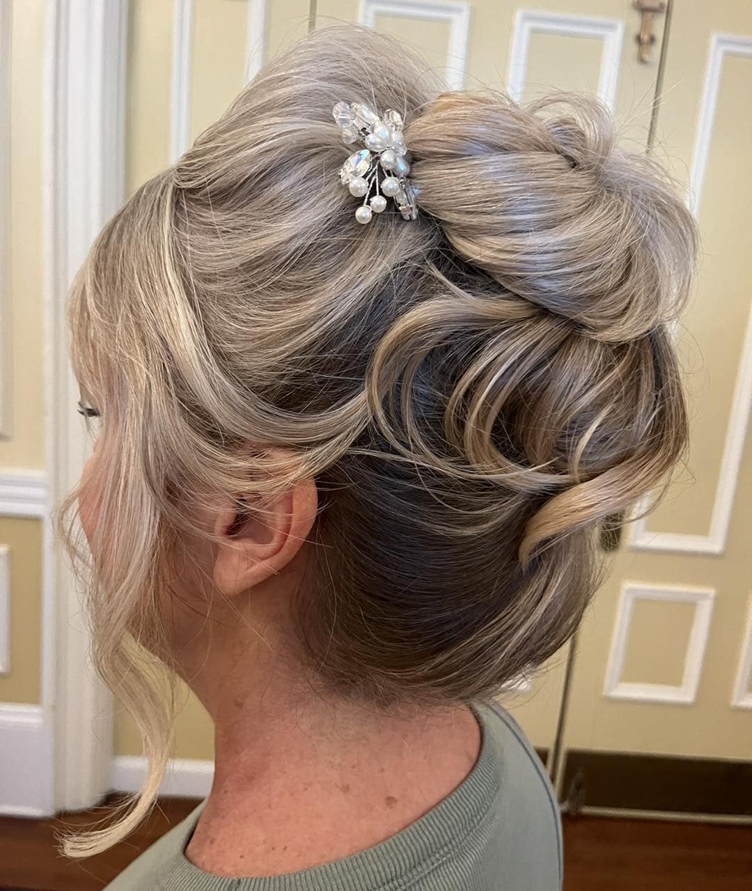 - Salone Estetica in Winchester, MA Woman with blonde hair color styled in an elegant updo, featuring loose curls and a pearl floral hair accessory, stands indoors against a cream-colored wall with decorative molding—perfect inspiration for wedding hair and makeup. - Salone Estetica in Winchester, MA