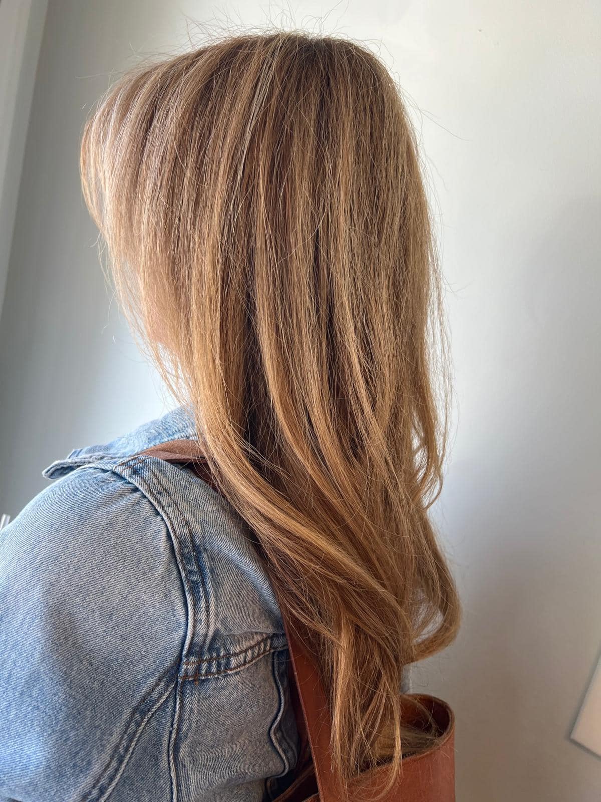 - Salone Estetica in Winchester, MA A person with long, light brown hair styled by a talented hair stylist, wearing a denim jacket and carrying a brown bag, faces away from the camera. - Salone Estetica in Winchester, MA