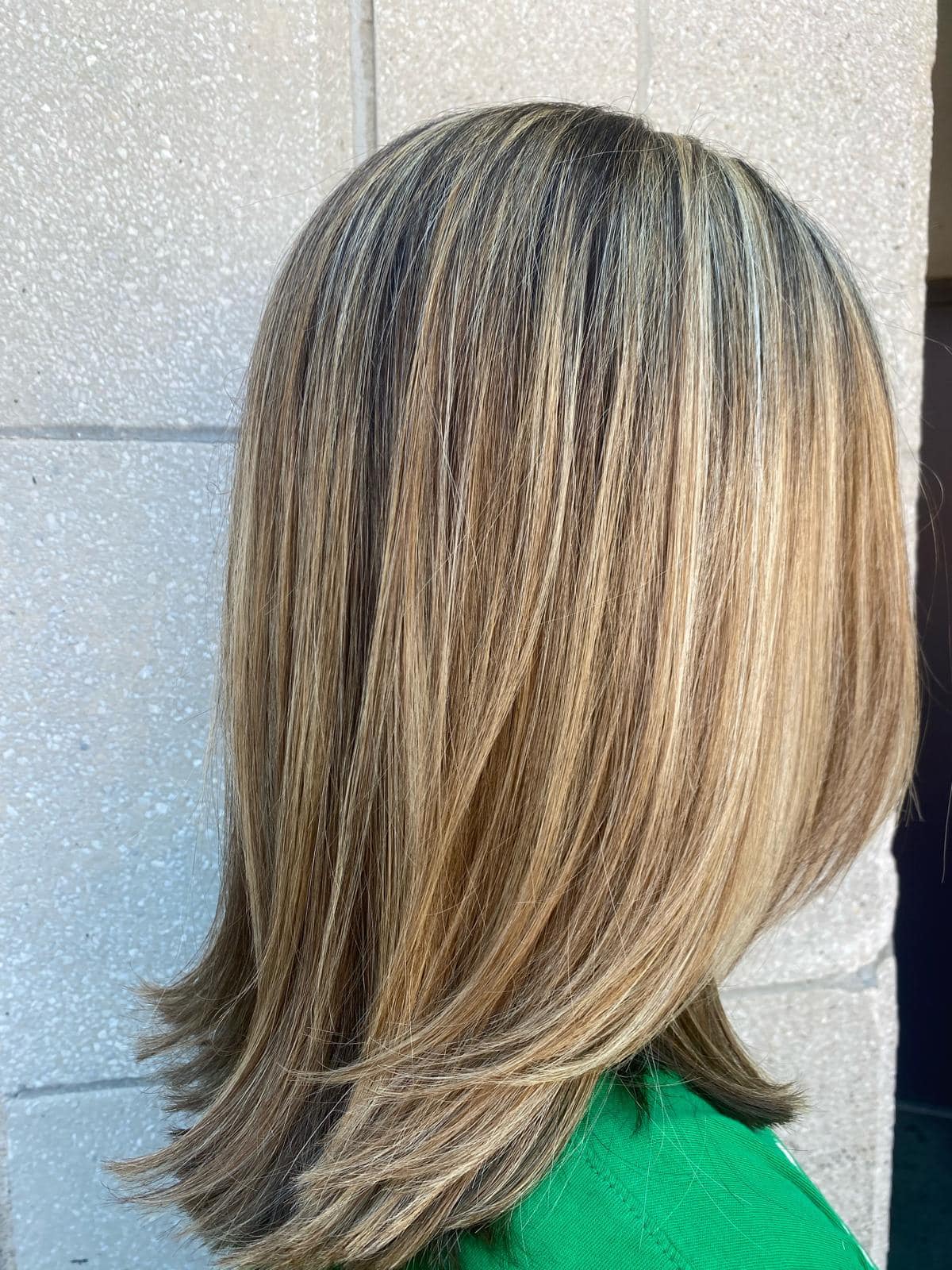 - Salone Estetica in Winchester, MA A person with straight, shoulder-length blonde hair with dark roots, standing against a light-colored textured wall—showcasing a stunning hair color that could come from the best salon or an expert Aveda salon. - Salone Estetica in Winchester, MA