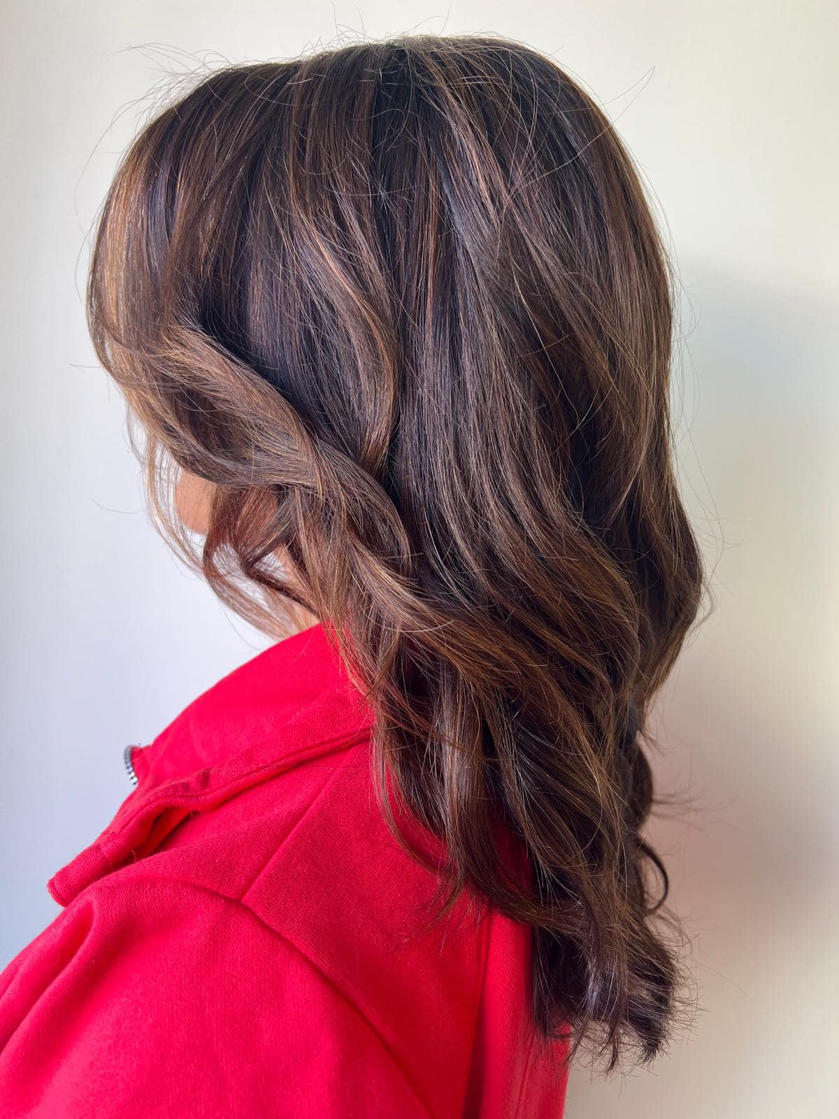 - Salone Estetica in Winchester, MA A person with wavy, medium-length brown hair color wearing a bright red top, shown from the side against a plain light background. - Salone Estetica in Winchester, MA