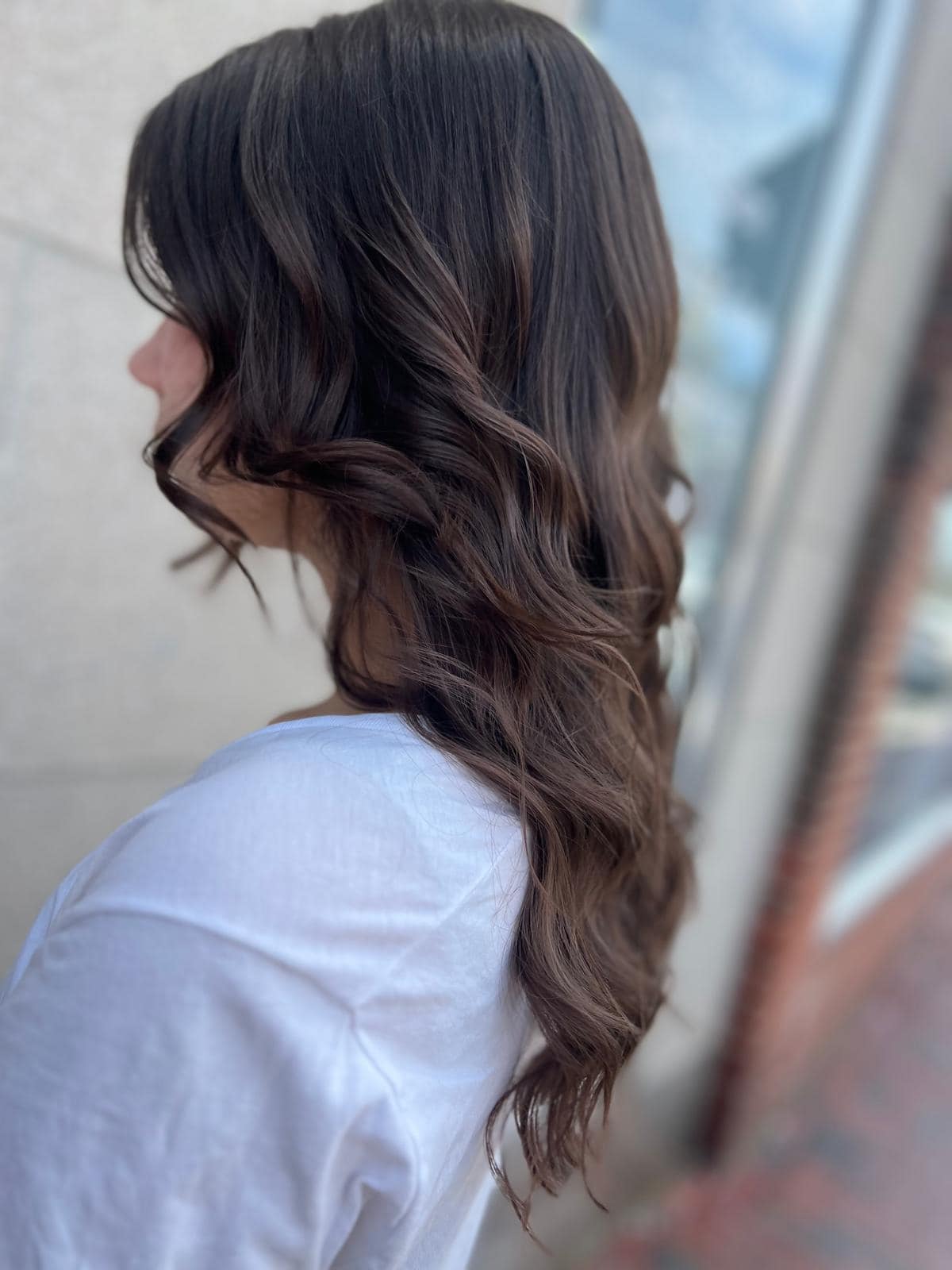 - Salone Estetica in Winchester, MA Person with long, wavy brown hair—styled to perfection by a professional hair stylist—wearing a white shirt is shown from the side, standing outdoors near a reflective window. - Salone Estetica in Winchester, MA