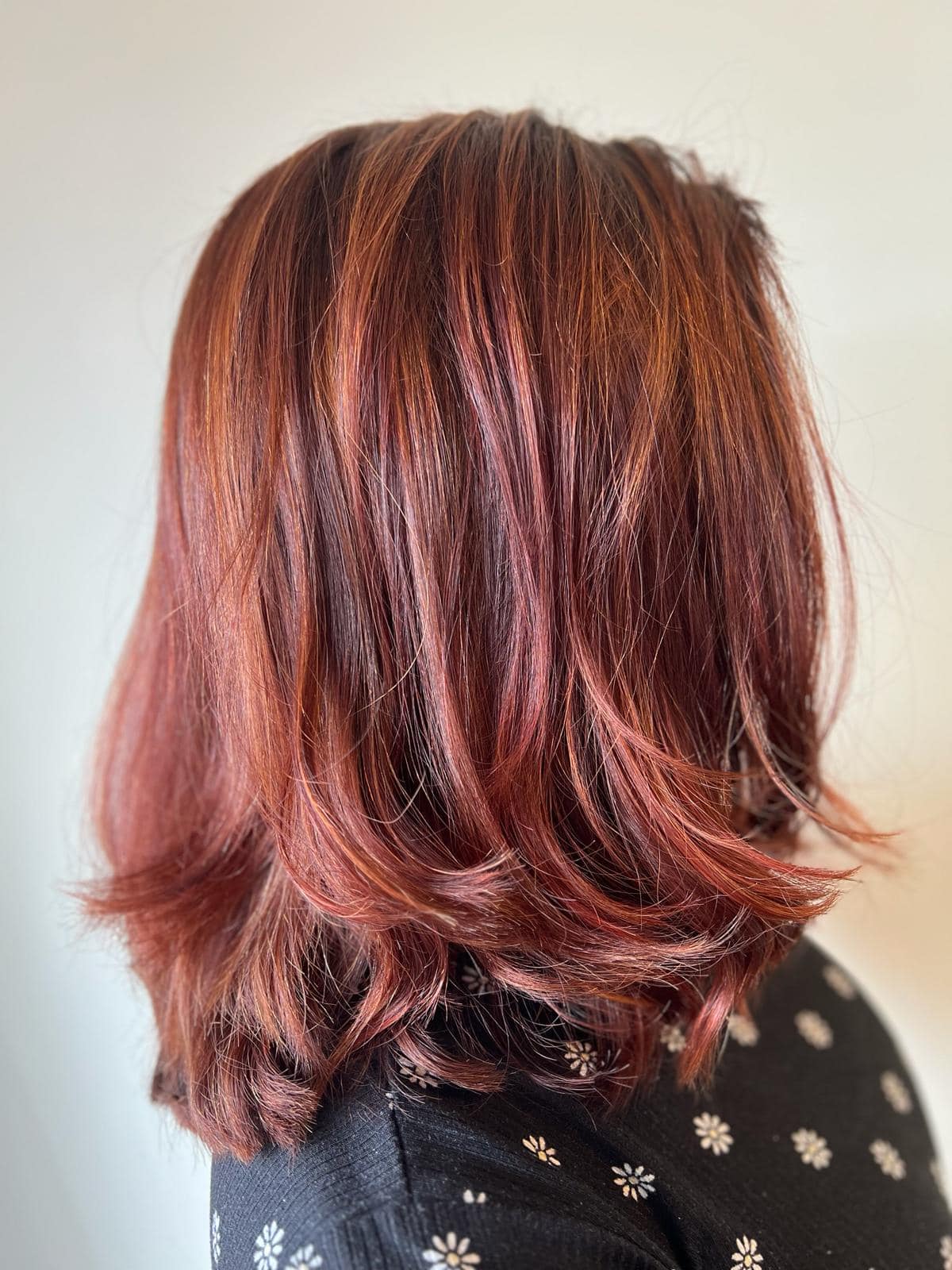 - Salone Estetica in Winchester, MA A person with shoulder-length, layered, reddish-brown hair—styled by a professional hair stylist—is photographed from the side, wearing a black top with a white floral pattern. - Salone Estetica in Winchester, MA